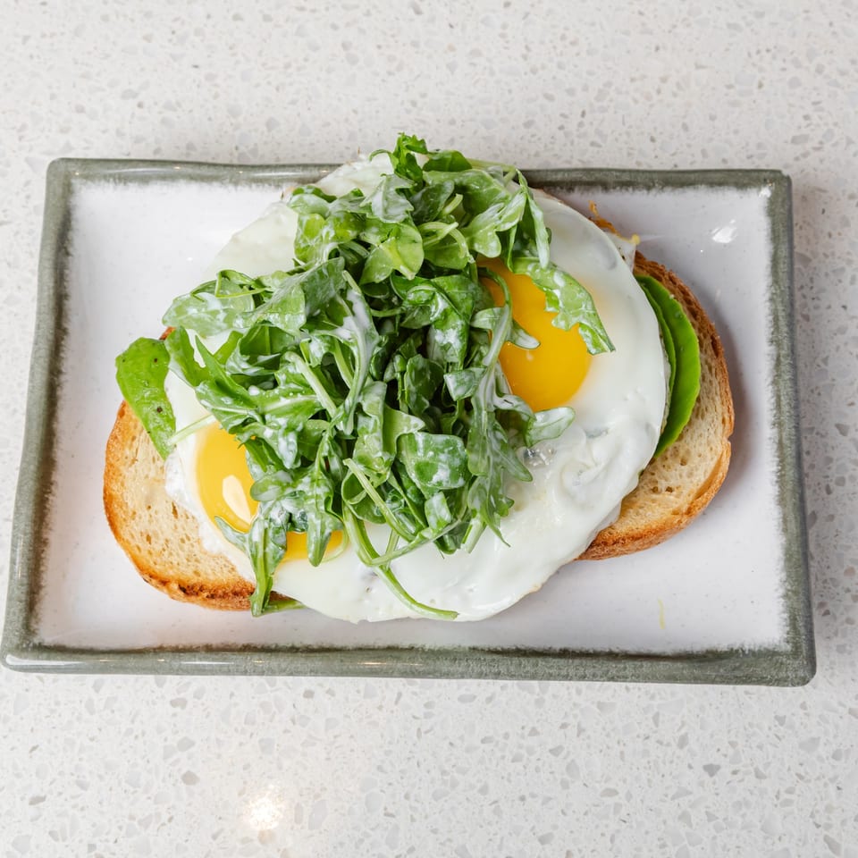 Toast topped with two fried eggs, avocado, and fresh arugula on a rectangular plate.