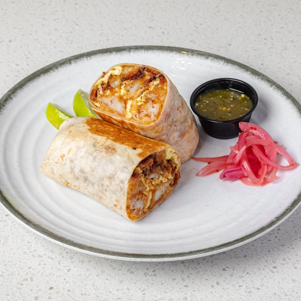 Two grilled, cut-open burritos with rice and fillings, served with lime wedges, pickled onions, and green salsa on a plate.