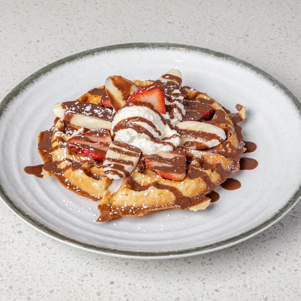 Belgian waffle topped with bananas, strawberries, whipped cream, chocolate sauce, and powdered sugar on a ceramic plate.
