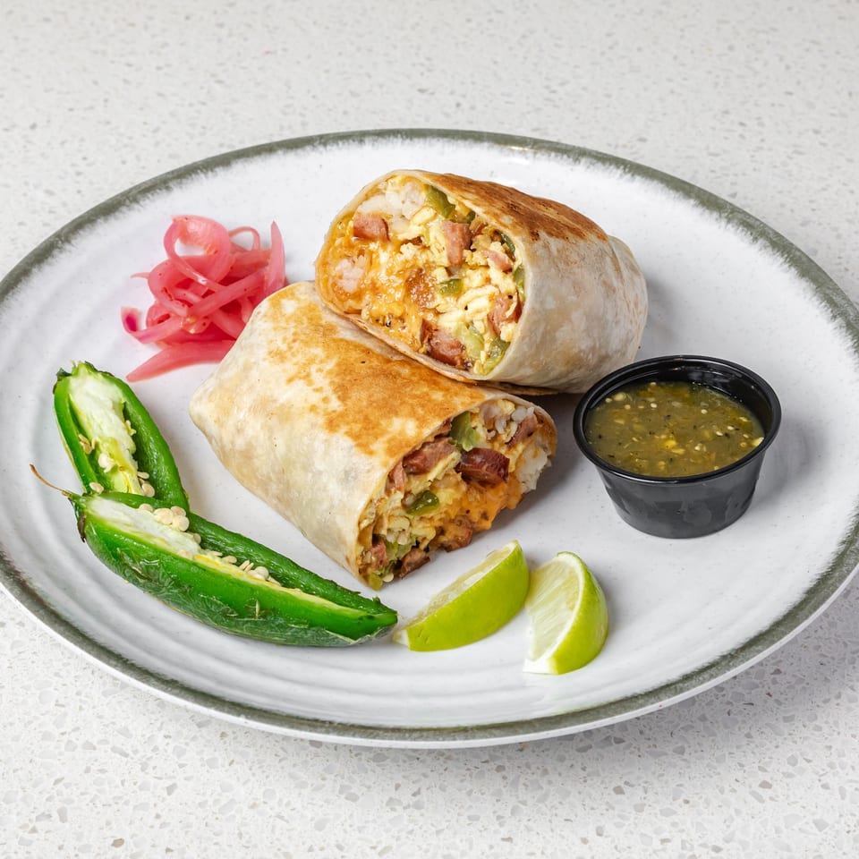 Two toasted breakfast burrito halves filled with eggs, sausage, cheese, and peppers, served with salsa, lime, jalapeño, and pickled onions.