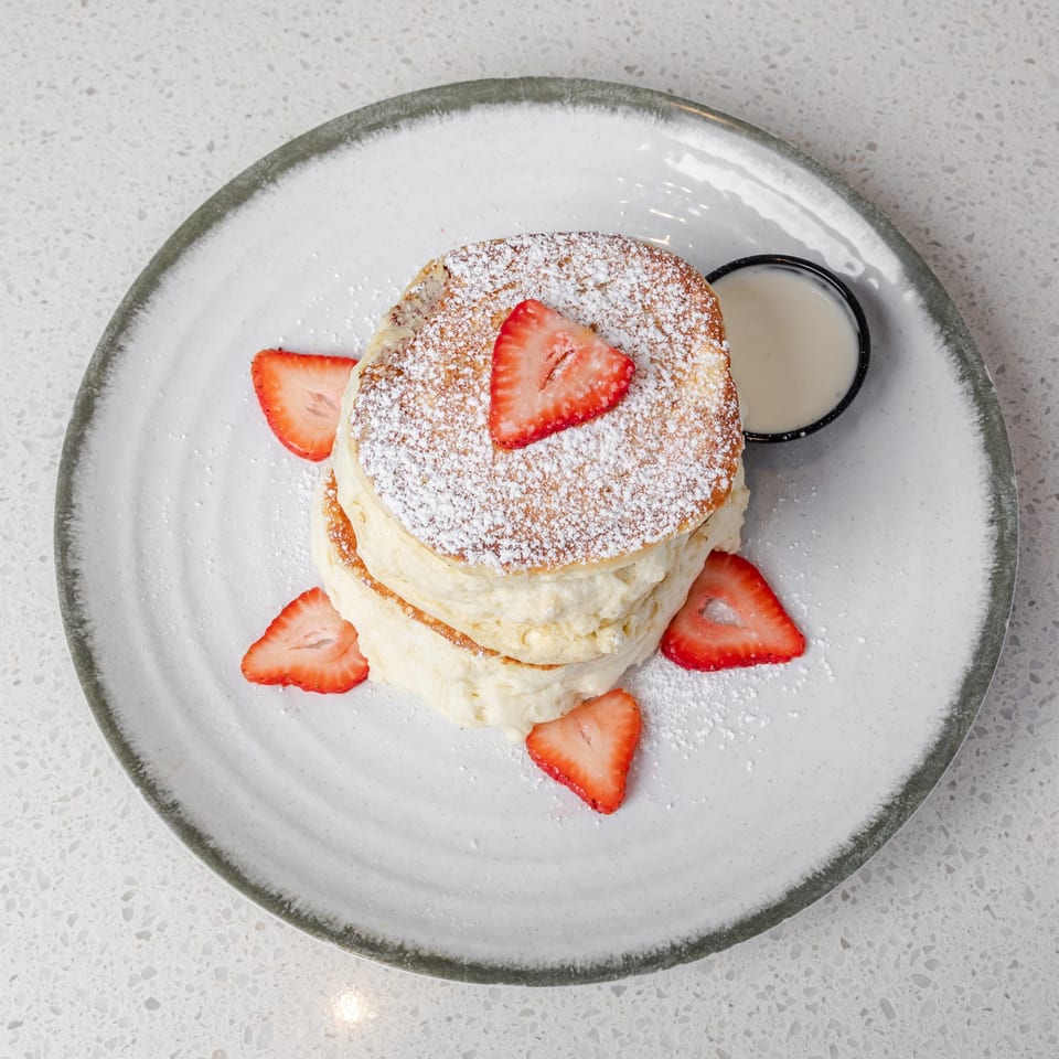 Stack of powdered sugar pancakes topped with strawberries, served with cream on a plate.