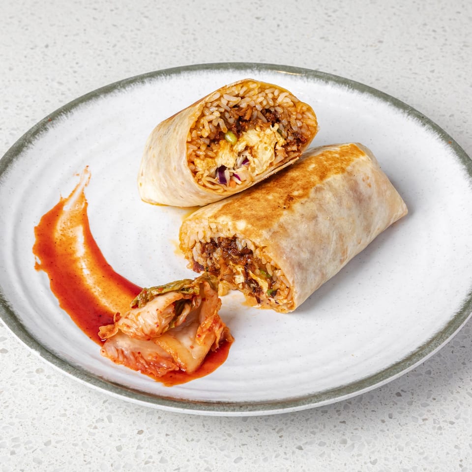Two toasted rice-filled burritos, one cut open with egg and vegetables, served with kimchi and red sauce on a plate.
