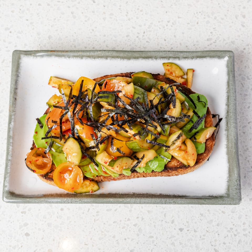 Open-faced toast topped with avocado, chopped tomatoes, cucumbers, lettuce, chili seasoning, and shredded nori on a rectangular plate.