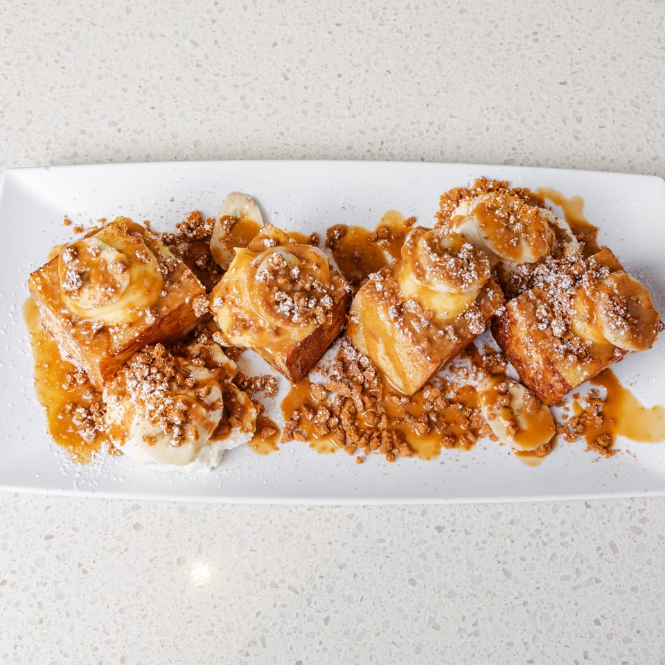 Caramel-drizzled cinnamon rolls topped with cream, cookie crumbles, and powdered sugar on a white rectangular plate.