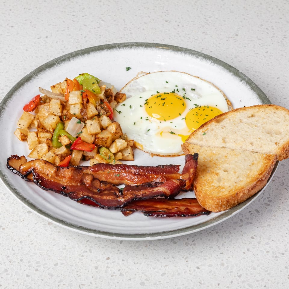 Breakfast plate with two fried eggs, roasted potatoes and peppers, crispy bacon, and toasted bread.