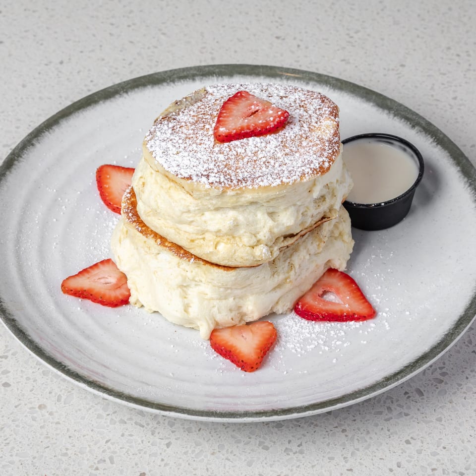 Stack of fluffy pancakes dusted with powdered sugar, topped with strawberries, and served with syrup on a plate.