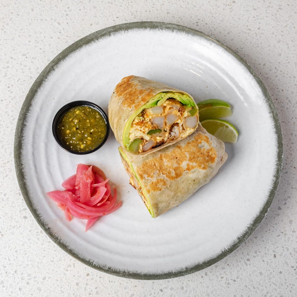Grilled chicken burrito cut in half, served with avocado, green salsa, lime wedges, and pickled onions on a plate.