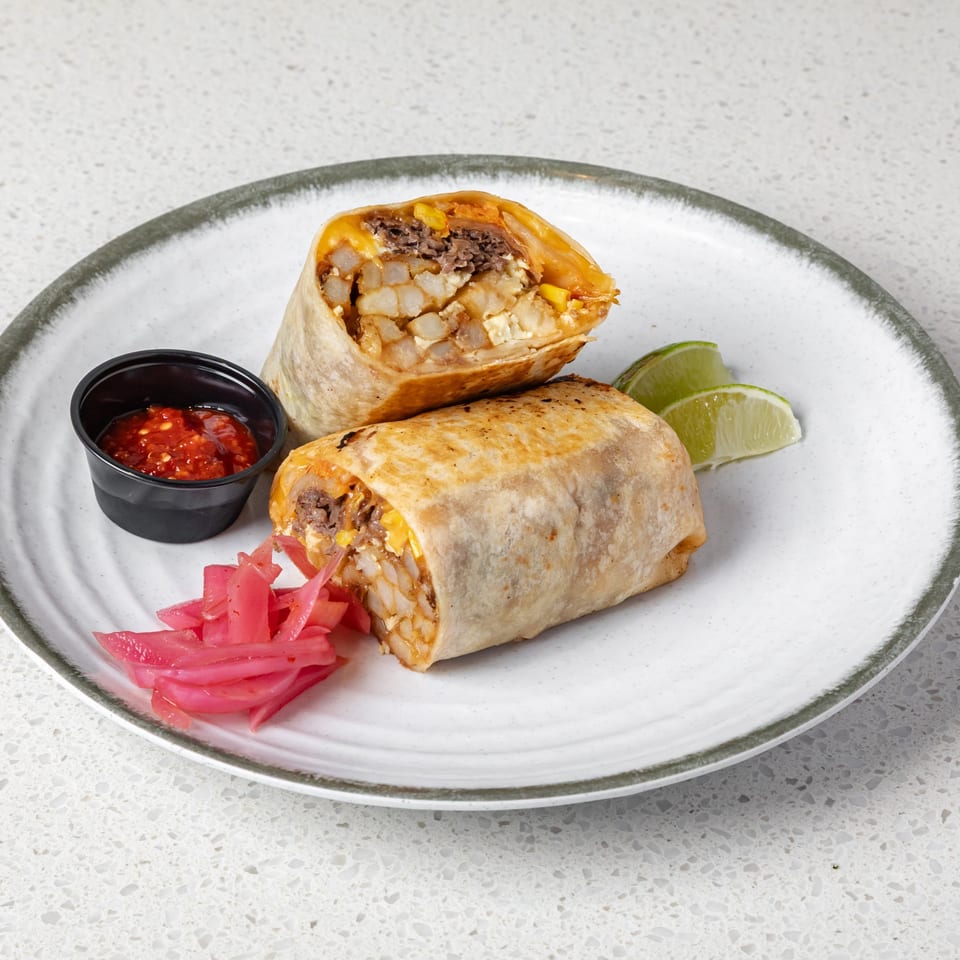 Two sliced beef and cheese burritos with potatoes, salsa, lime wedges, and pickled onions on a plate.