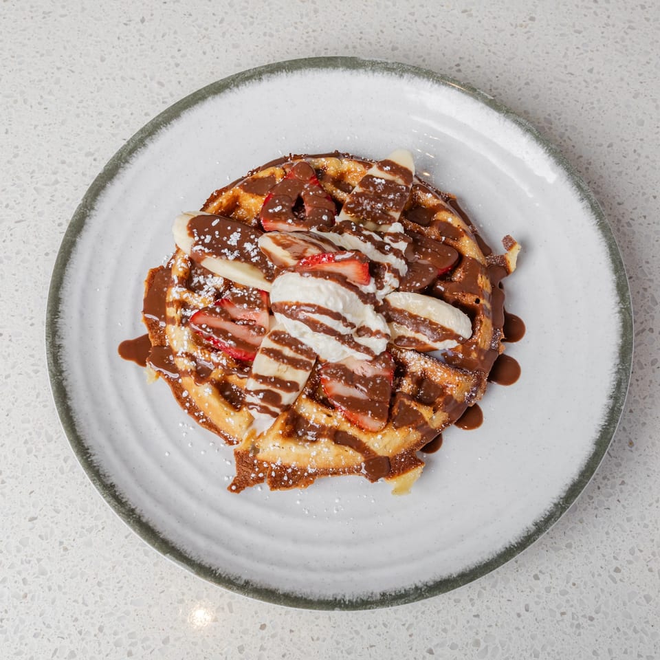 Golden waffle topped with sliced bananas, strawberries, whipped cream, chocolate sauce, and powdered sugar on a ceramic plate.