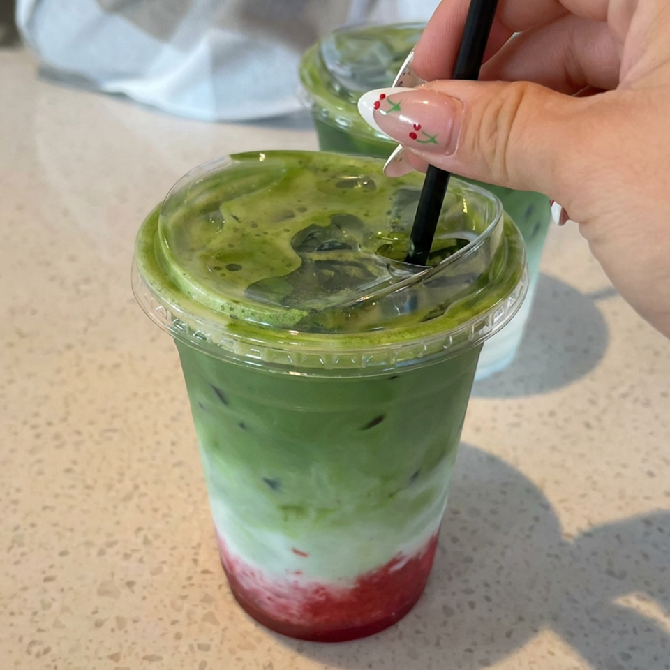Hand stirs an iced green matcha drink layered with milk and red fruit in a clear plastic cup.