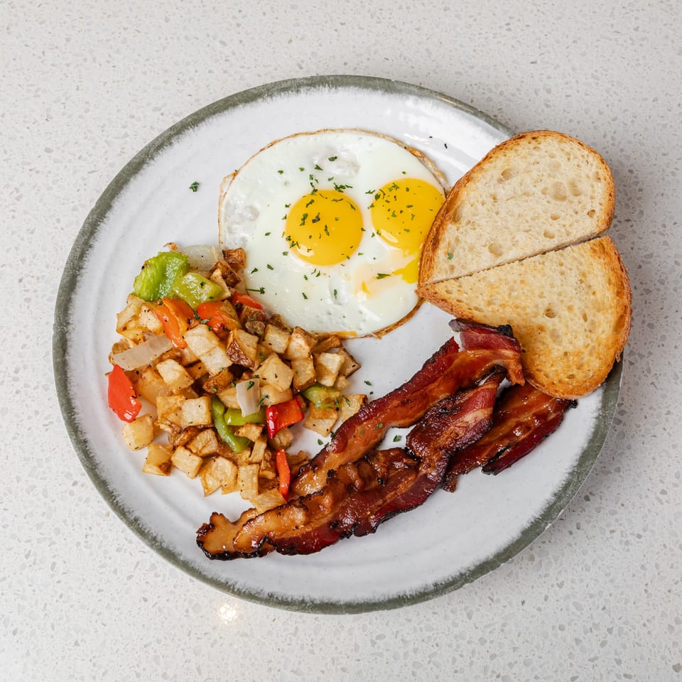 Breakfast plate with two fried eggs, roasted potatoes and peppers, bacon, and toasted bread.