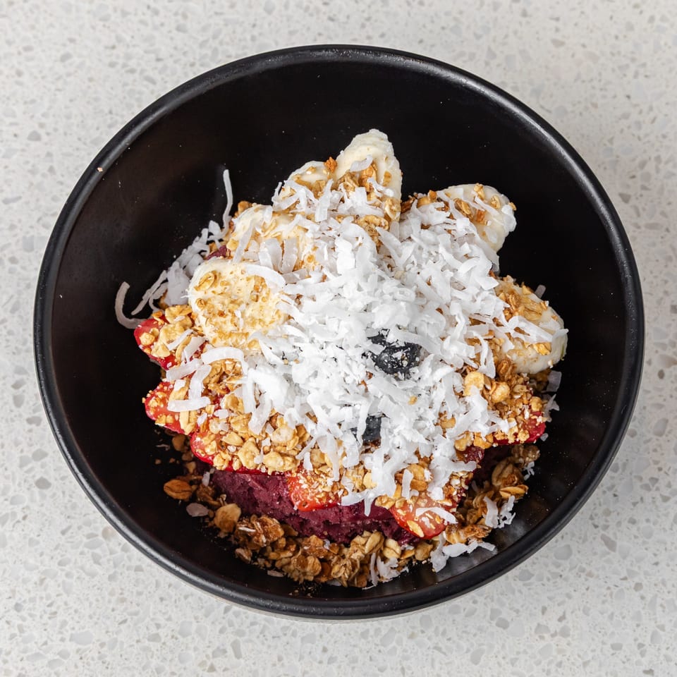 Açaí bowl topped with sliced bananas, strawberries, granola, shredded coconut, and blueberries in a black bowl.